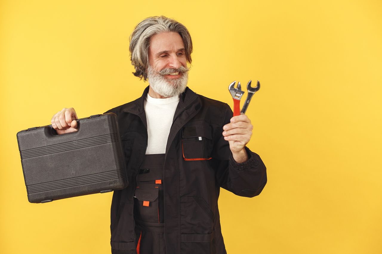 Services Elderly bearded man with safety glasses smiling, holding a toolbox and wrenches against a yellow background.