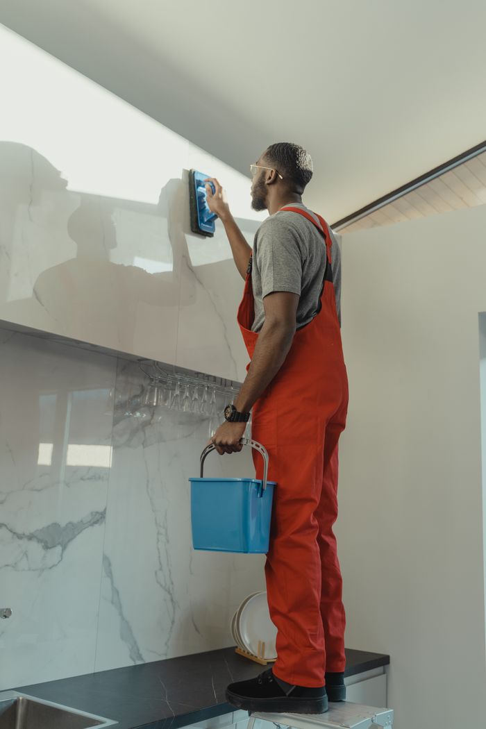 Services African American man in red overalls cleaning a kitchen wall with a brush and pail.