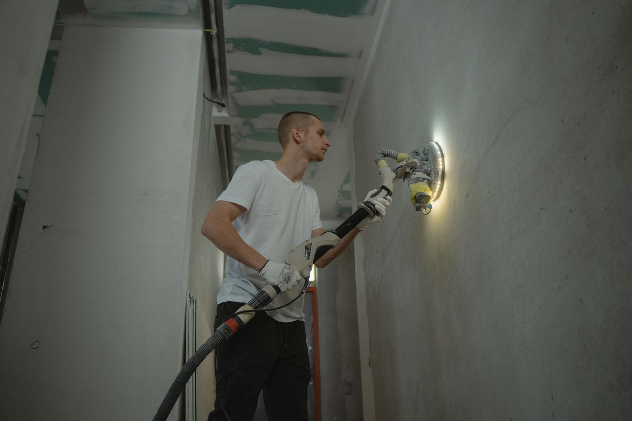 A man sanding an interior wall with an automatic sanding tool during renovation work indoors.